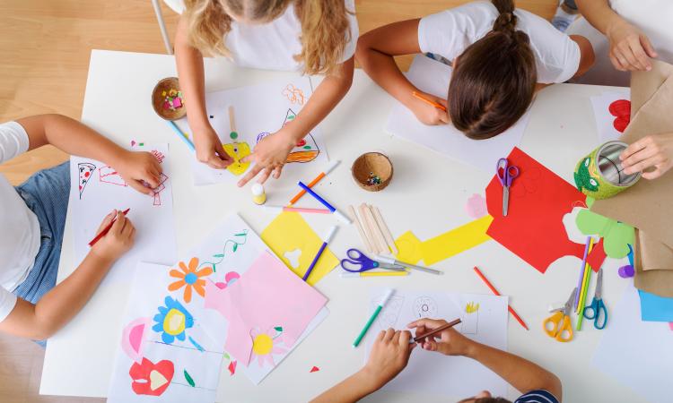An overhead photo of a children's arts and crafts sessions. There's brightly-coloured paper, scissors and felt tip pens on the table with the children around it on all sides